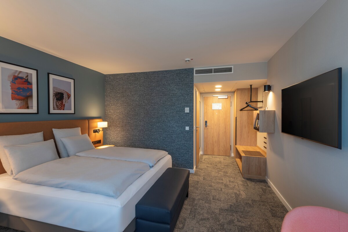 A hotel room with two beds, a bench, a TV, and artwork on the walls.