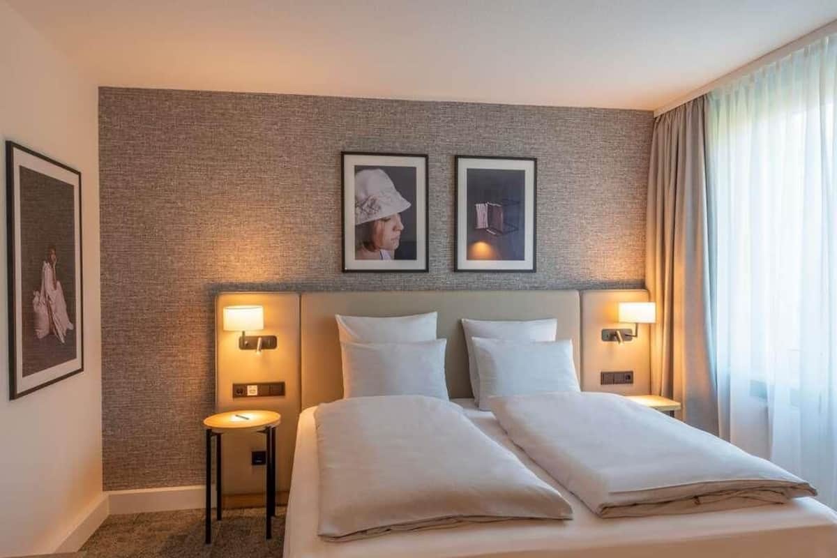 A hotel room with a bed, two bedside lamps, a small table, and two framed pictures on the wall.