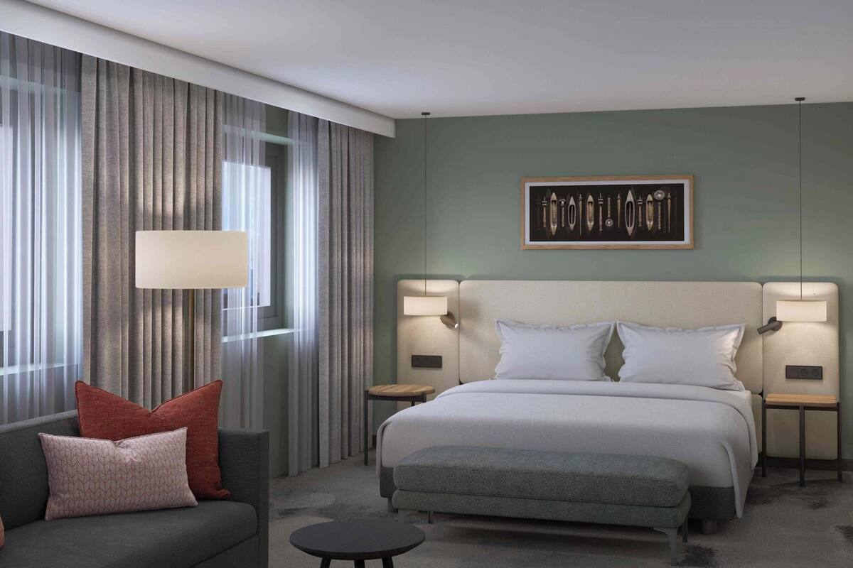 A modern hotel room with a large bed, two bedside tables with lamps, a sofa with cushions, a small round table, and a large window with curtains.