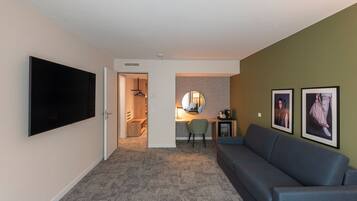 Habitación de hotel con televisor de pantalla plana, sofá, escritorio con silla y obra de arte en la pared.