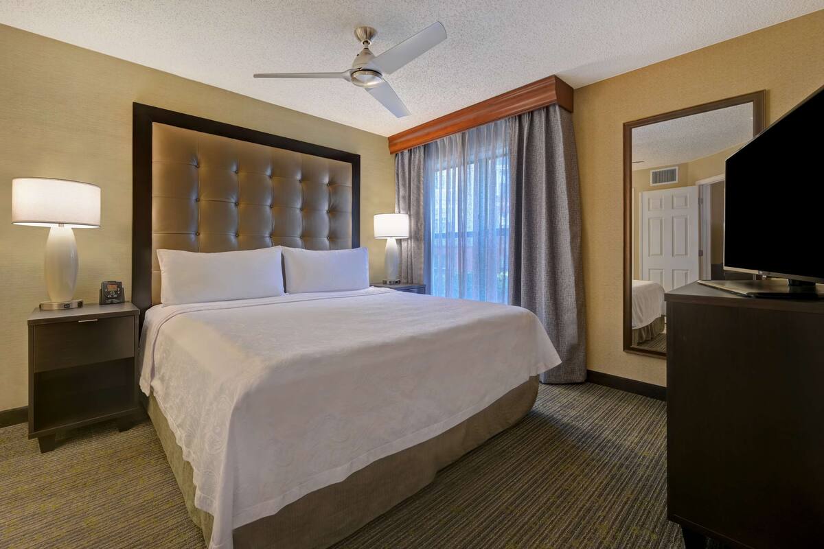 A hotel room with a large bed, two bedside tables with lamps, a ceiling fan, a television, and a large mirror.