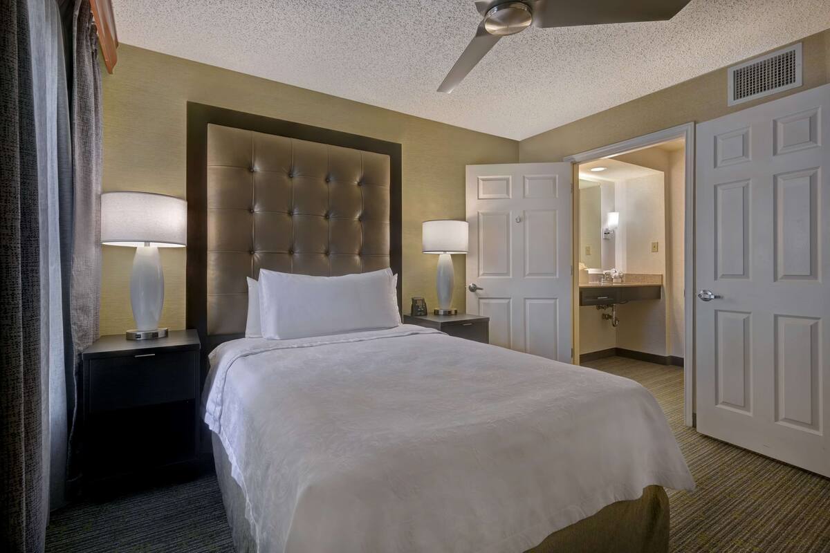 A hotel room with a large bed, two bedside lamps, a ceiling fan, and a bathroom visible through an open door.