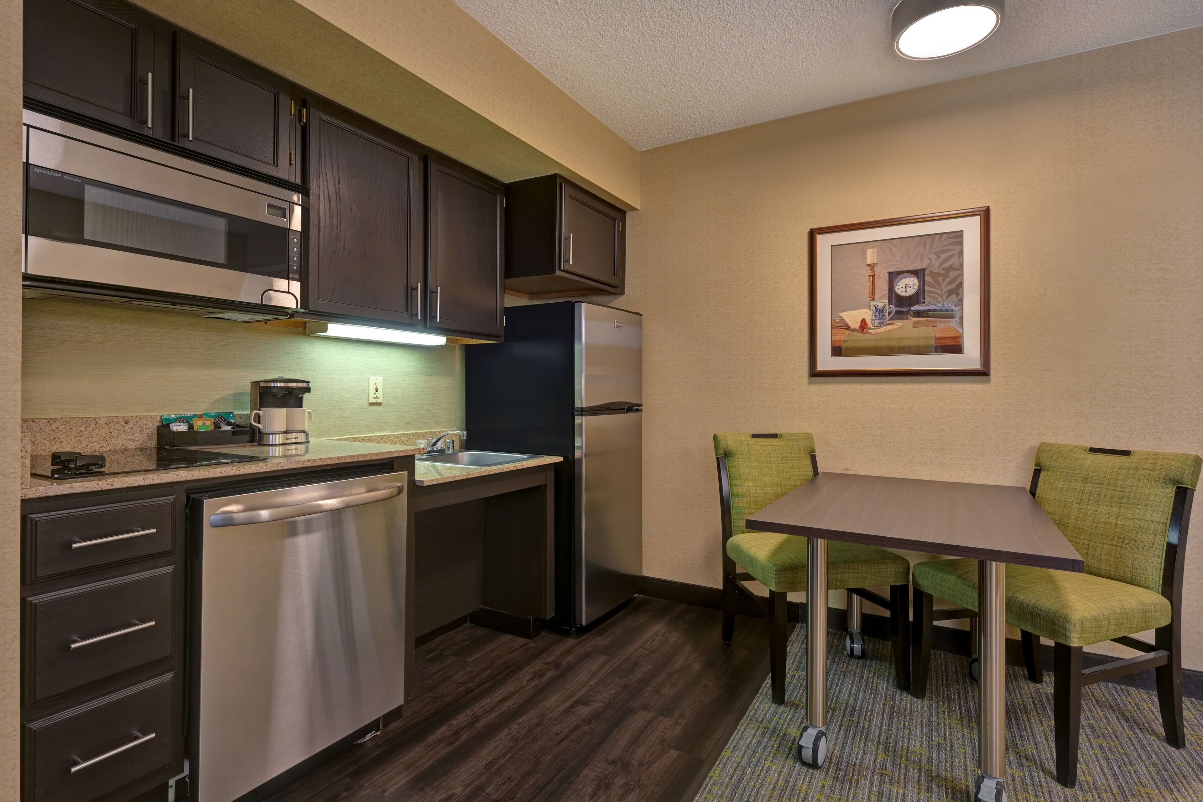 A compact kitchen with dark cabinets, a stainless steel dishwasher, and a refrigerator. There is a small dining area with a table and two chairs.