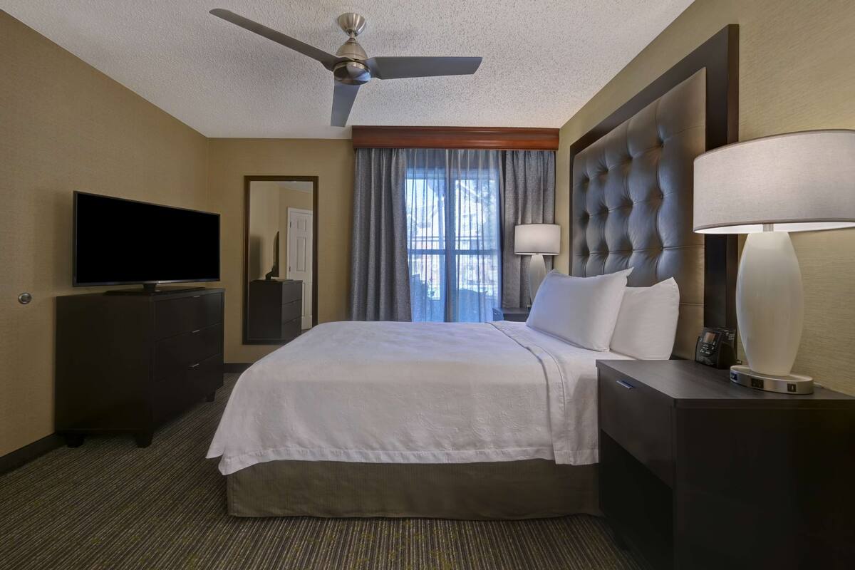 A hotel room with a bed, a TV, a desk with a lamp, a ceiling fan, and a window with curtains.