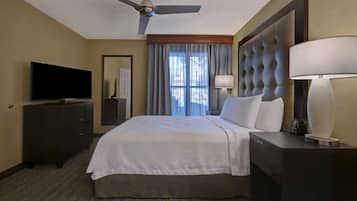 A hotel room with a bed, a TV, a desk with a lamp, a ceiling fan, and a window with curtains.