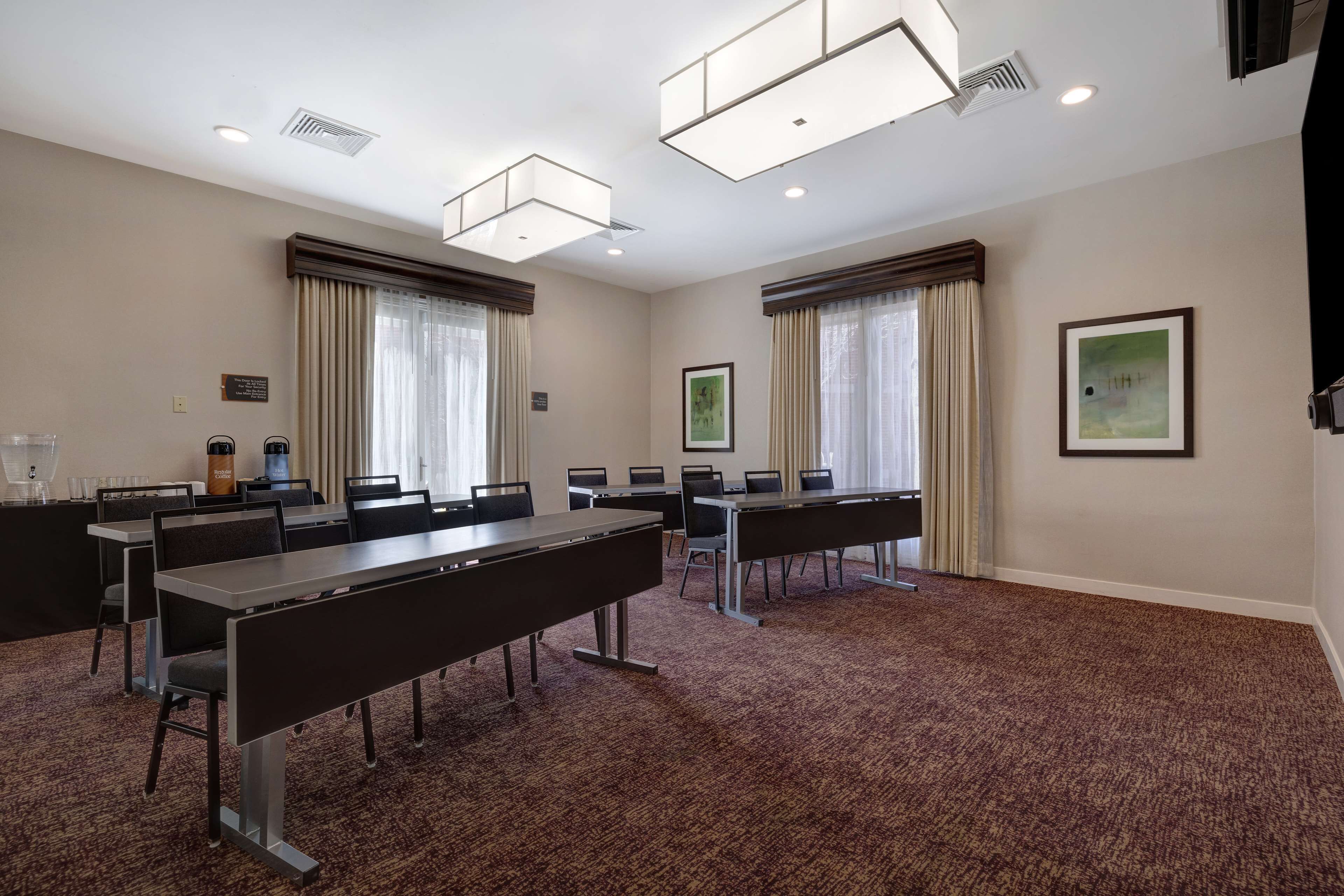 A conference room with multiple tables and chairs, large windows with curtains, and ceiling lights.