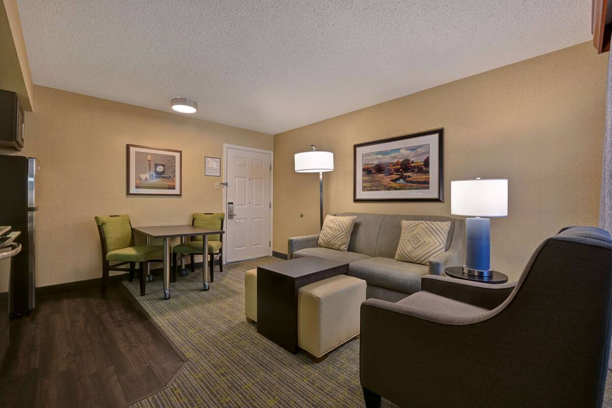 A hotel room with a sofa, ottoman, dining table with chairs, a lamp, and a television.
