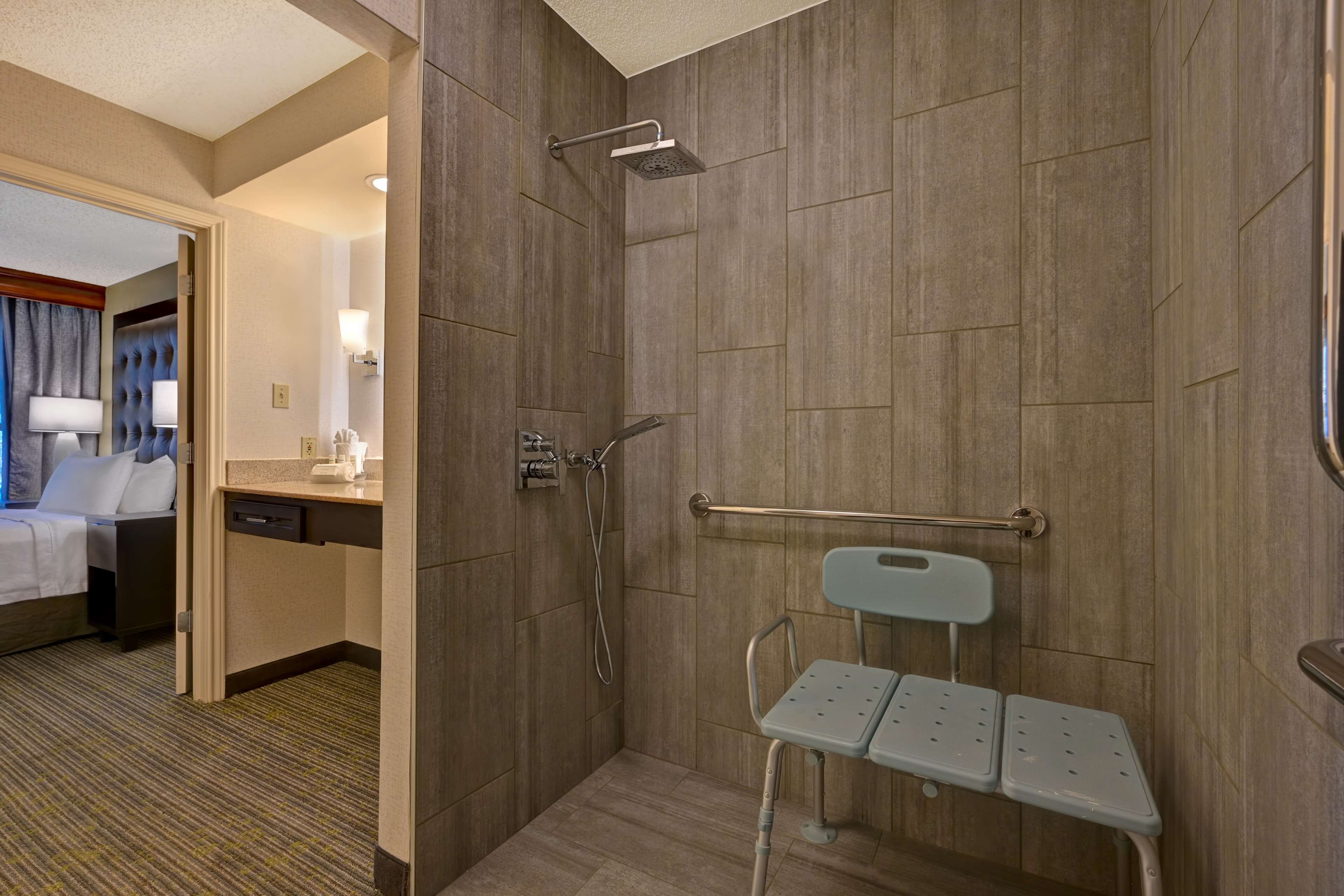 A hotel bathroom with a shower, a toilet with a grab bar, and a small seating area.