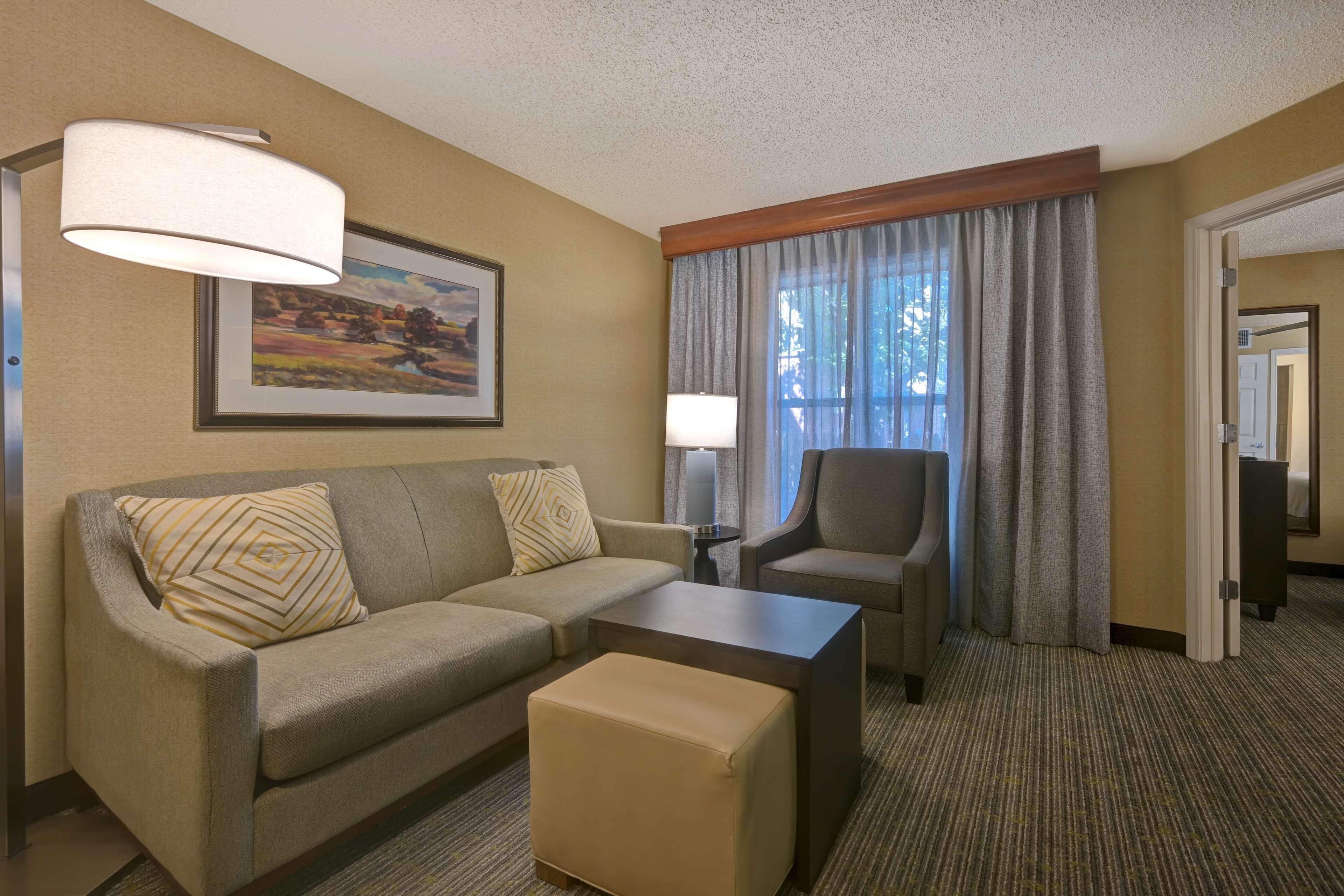 A hotel room with a sofa, ottoman, armchair, and a lamp. There is a painting on the wall and a window with curtains.