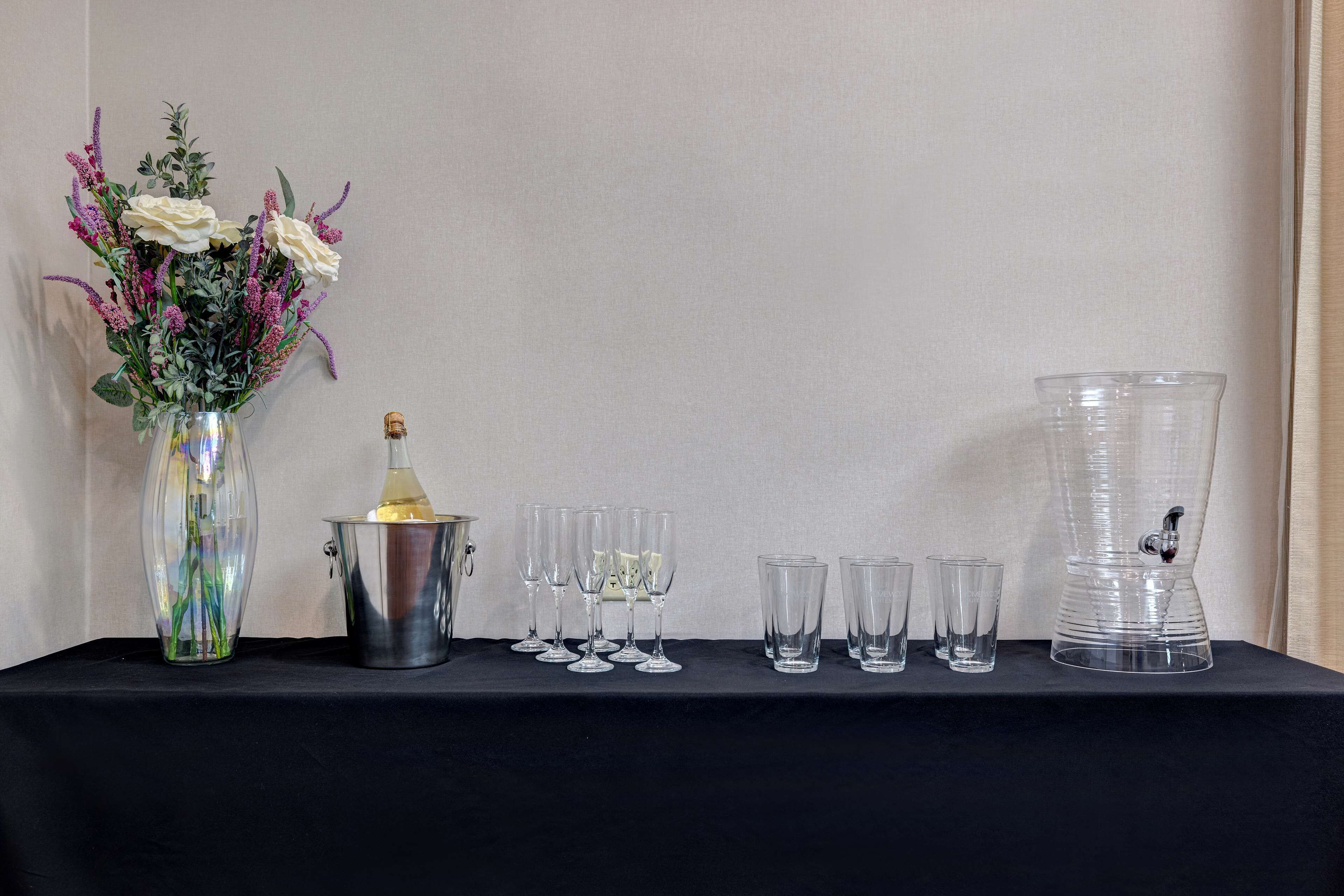 A table with a vase of flowers, a bottle, and several glasses and a water dispenser.