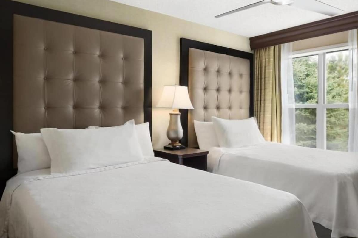A hotel room with two beds, a nightstand with a lamp, and a window with curtains.