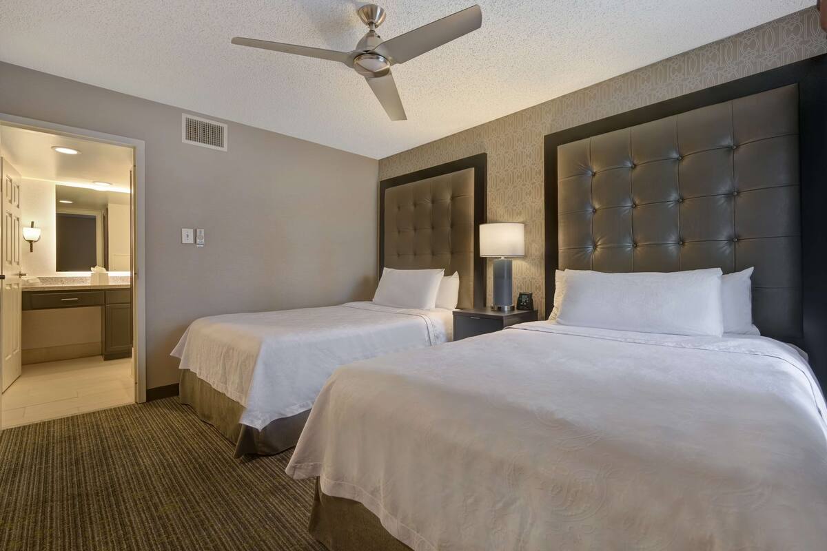A hotel room with two beds, a ceiling fan, a nightstand with a lamp, and a bathroom visible through an open door.