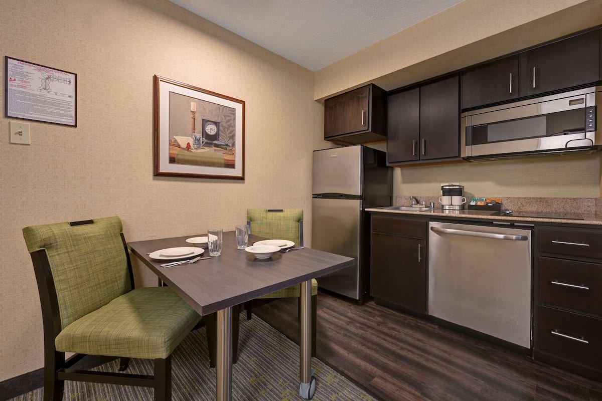 A compact hotel room with a dining table, two chairs, a refrigerator, a microwave, a dishwasher, and a framed picture on the wall.