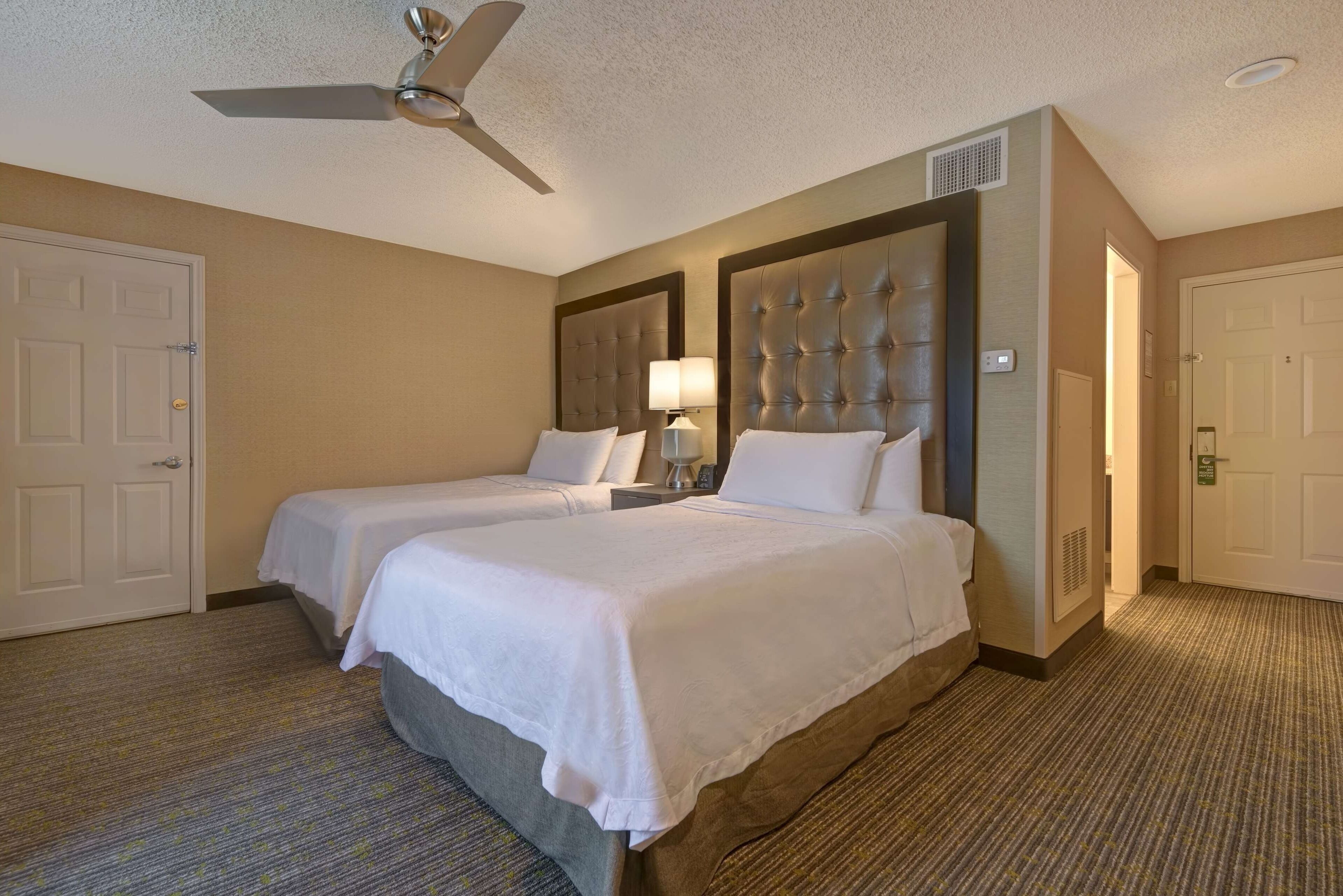 A hotel room with two beds, a ceiling fan, a lamp, and a carpeted floor.