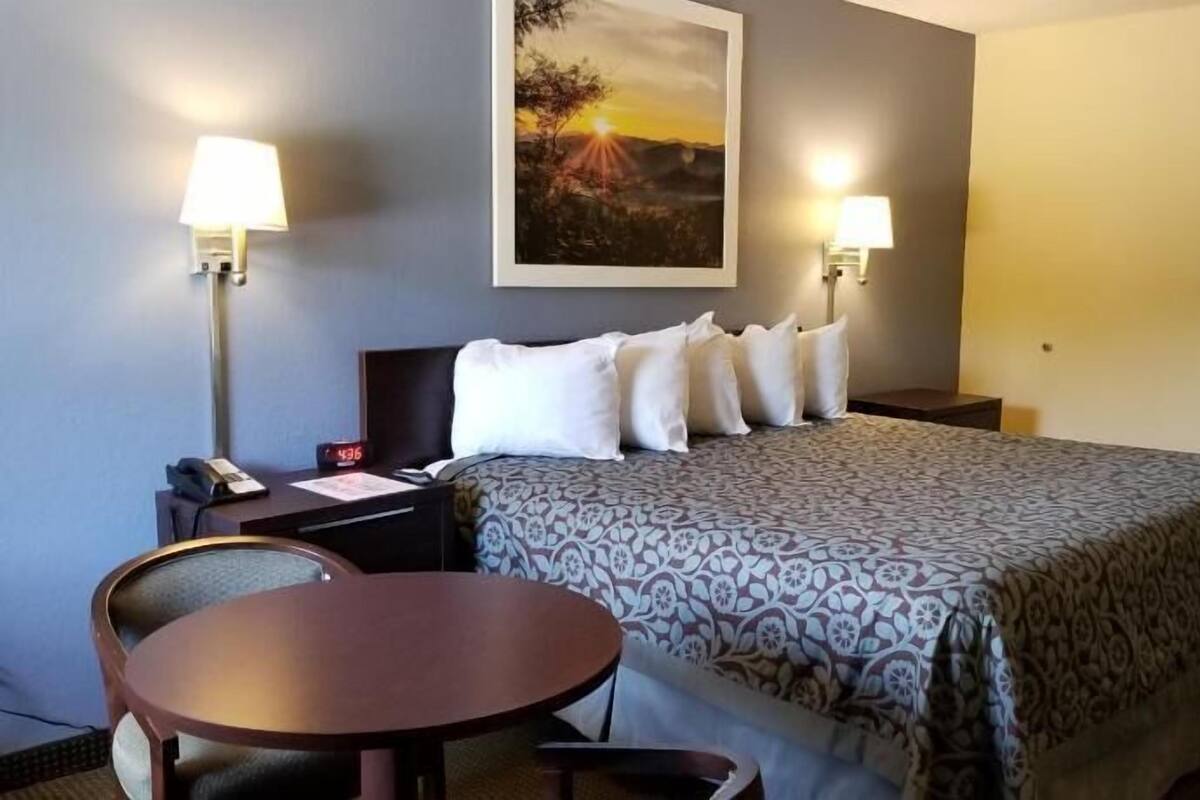 A hotel room with a bed, two bedside lamps, a painting, a desk with a chair, and a door.