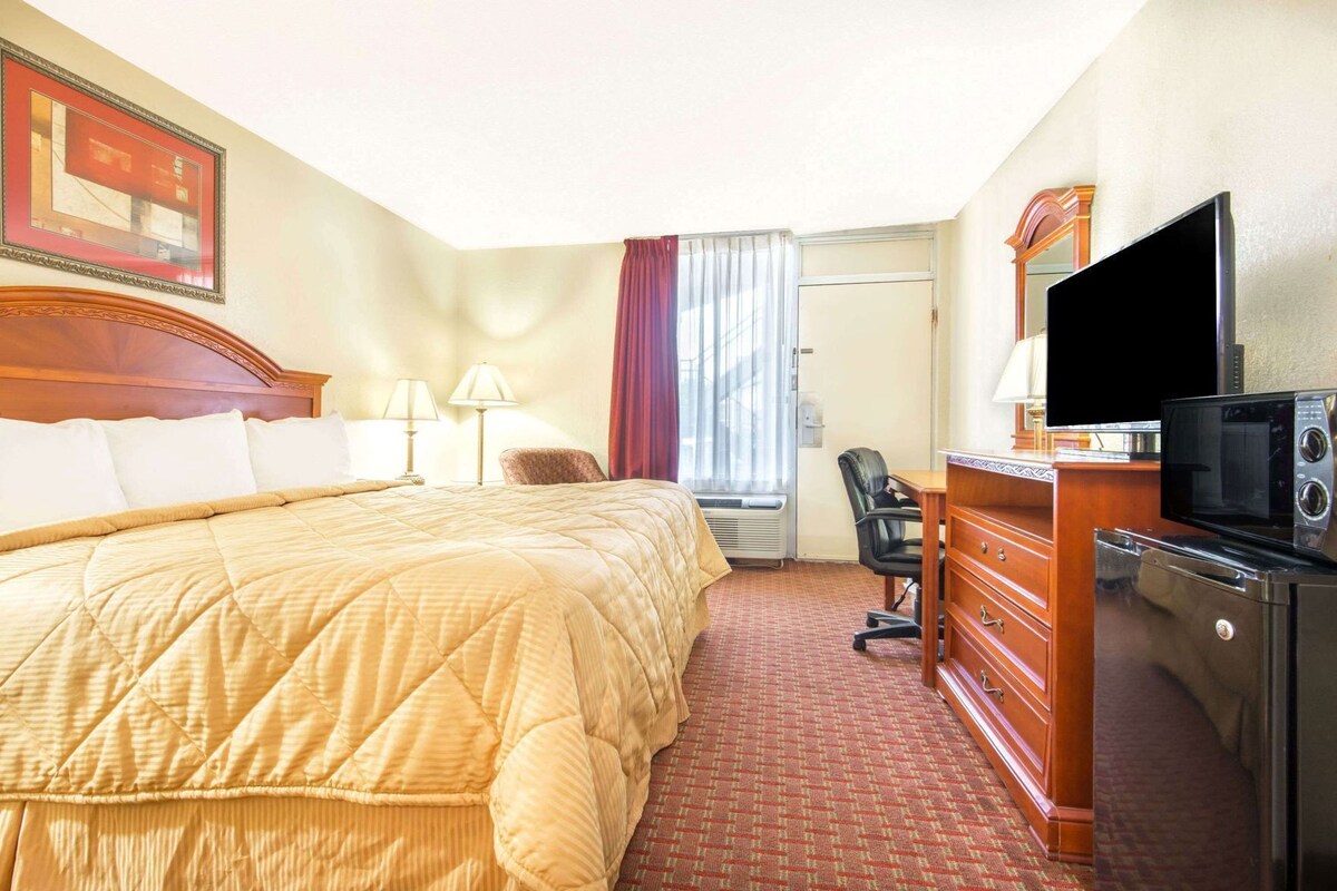 A hotel room with a large bed, a desk with a television and microwave, a chair, a dresser, and a window with curtains.