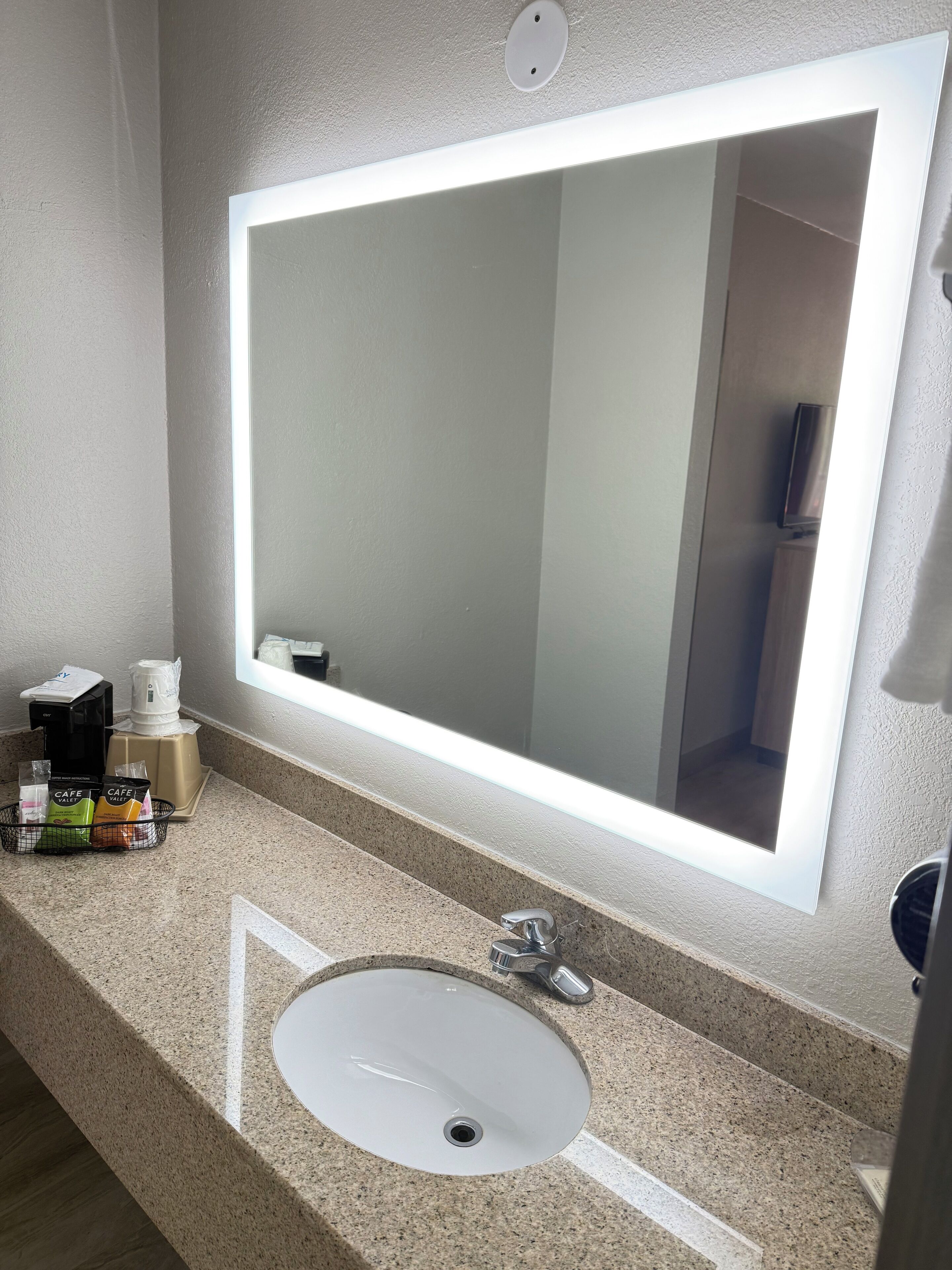 Ein Badezimmer mit einem großen Spiegel, einem Waschbecken und einer Kaffeemaschine.