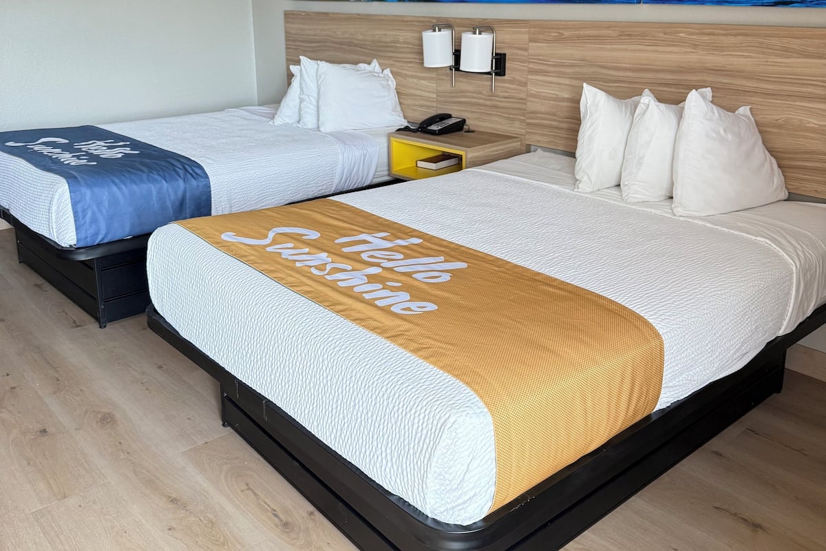 Ett hotellrum med två sängar, en sänggavel i trä, en tavla som föreställer en solnedgång och en kudde med texten ”Hej, solsken!”.