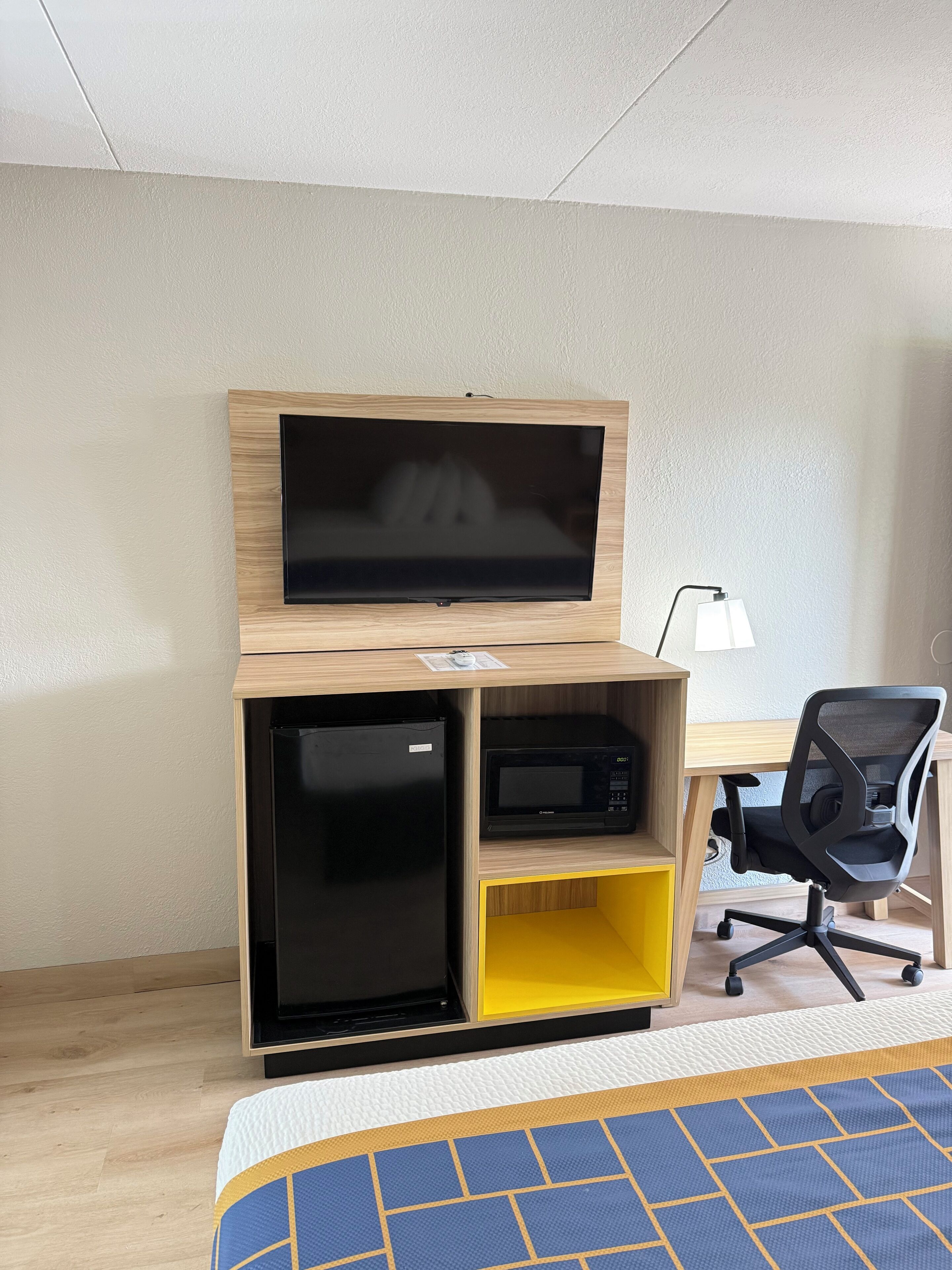 A compact room with a bed, a desk with a flat-screen TV, a microwave, and a chair.