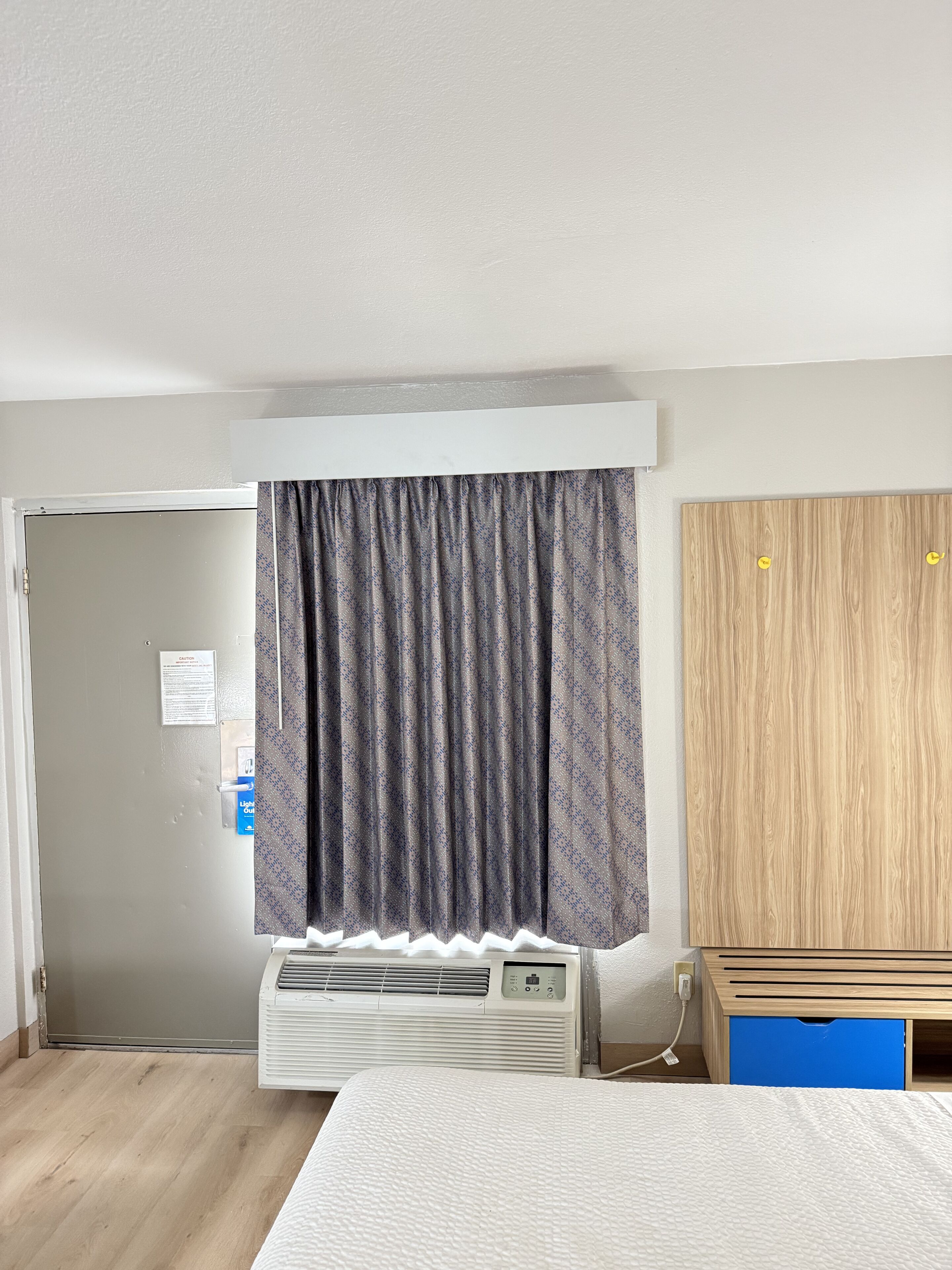 Habitació d'hotel amb un llit, un aire condicionat, un armari de fusta, un moble blau i una porta amb un senyal de precaució.
