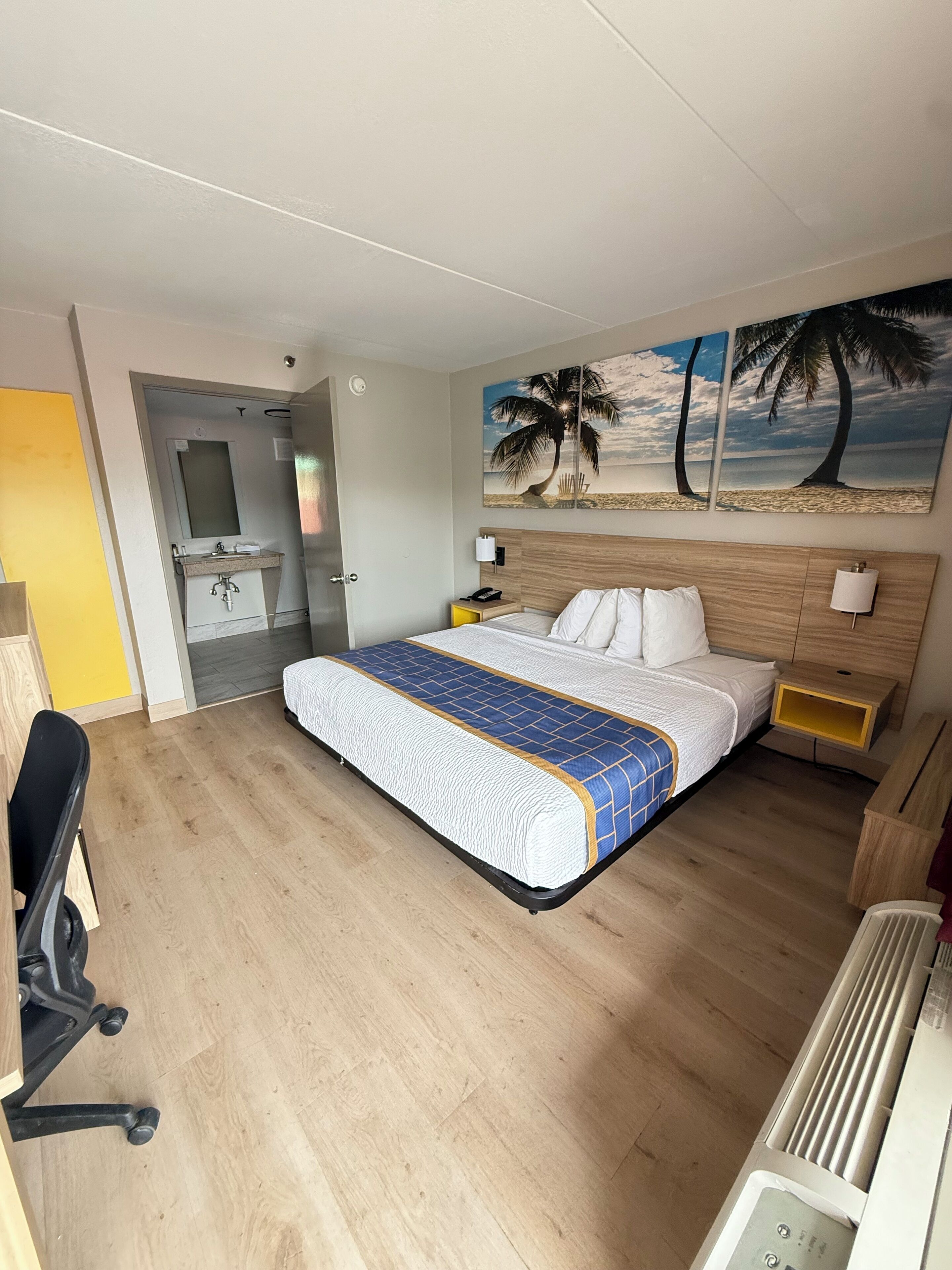 Una habitación de hotel con una cama, un escritorio, una silla, una mesita de noche, una lámpara de pared y un cuadro de una playa.