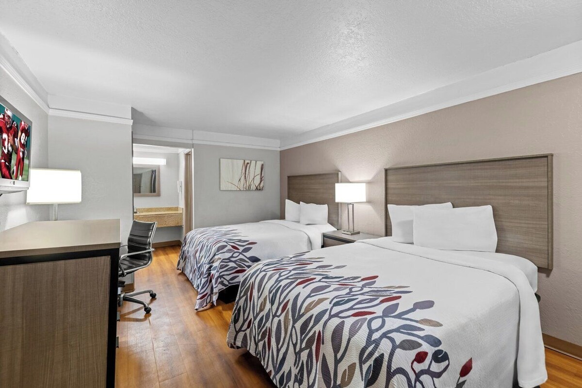 A hotel room with two beds, a desk, a chair, a TV, and a bathroom visible through an open door.