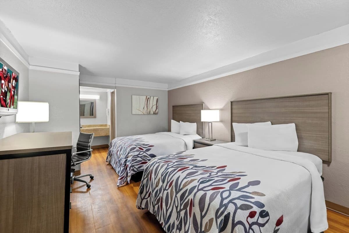 A hotel room with two beds, a desk, a chair, a TV, and a bathroom visible through an open door.