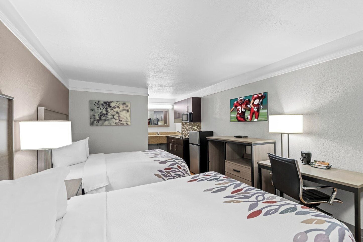 A hotel room with two beds, a desk, a chair, a TV, and a bathroom visible in the background.