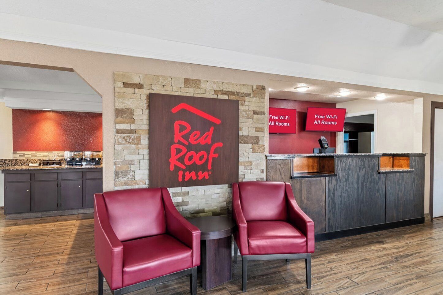 L'aire d'accueil d'un hôtel Red Roof Inn, avec un mur en pierre, un panneau indicateur, un comptoir de réception et deux fauteuils en cuir rouge.