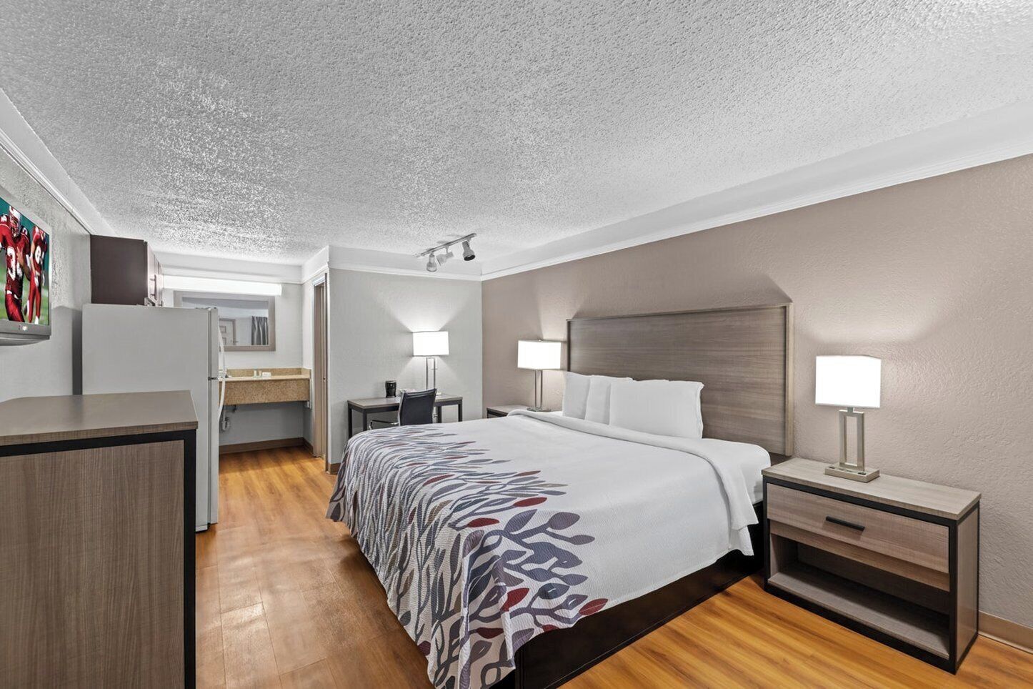 A hotel room with a bed, bedside tables, a desk, a chair, a mirror, and a kitchenette.