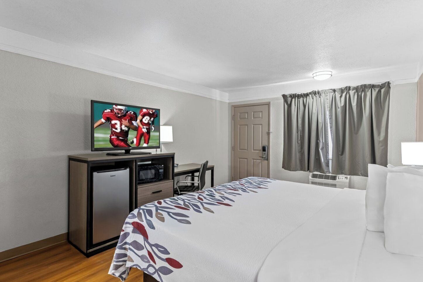 Quarto de hotel com cama, escrivaninha, televisão, frigorífico, cadeira e janela com cortinas.