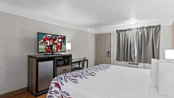 Quarto de hotel com cama, escrivaninha, televisão, frigorífico, cadeira e janela com cortinas.