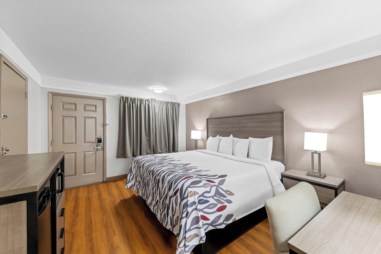 A hotel room with a bed, bedside table, lamp, desk, chair, wardrobe, and a door.