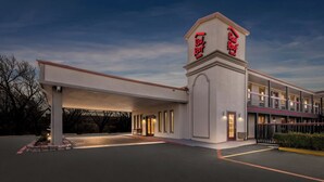 Ein Motel mit einem zentralen Turm, der das Red Roof Inn Logo zeigt, einem überdachten Parkbereich und einem mehrstöckigen Gebäude mit Balkonen.