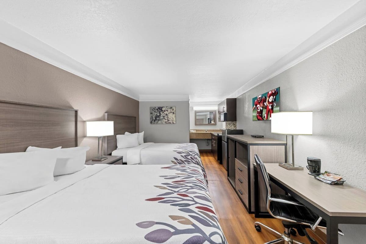 A hotel room with a large bed, a desk, a chair, a TV, and a kitchenette.