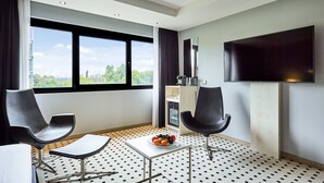 A modern hotel room with two black chairs, a small table with a fruit bowl, a flat-screen TV, and a large window offering a city view.