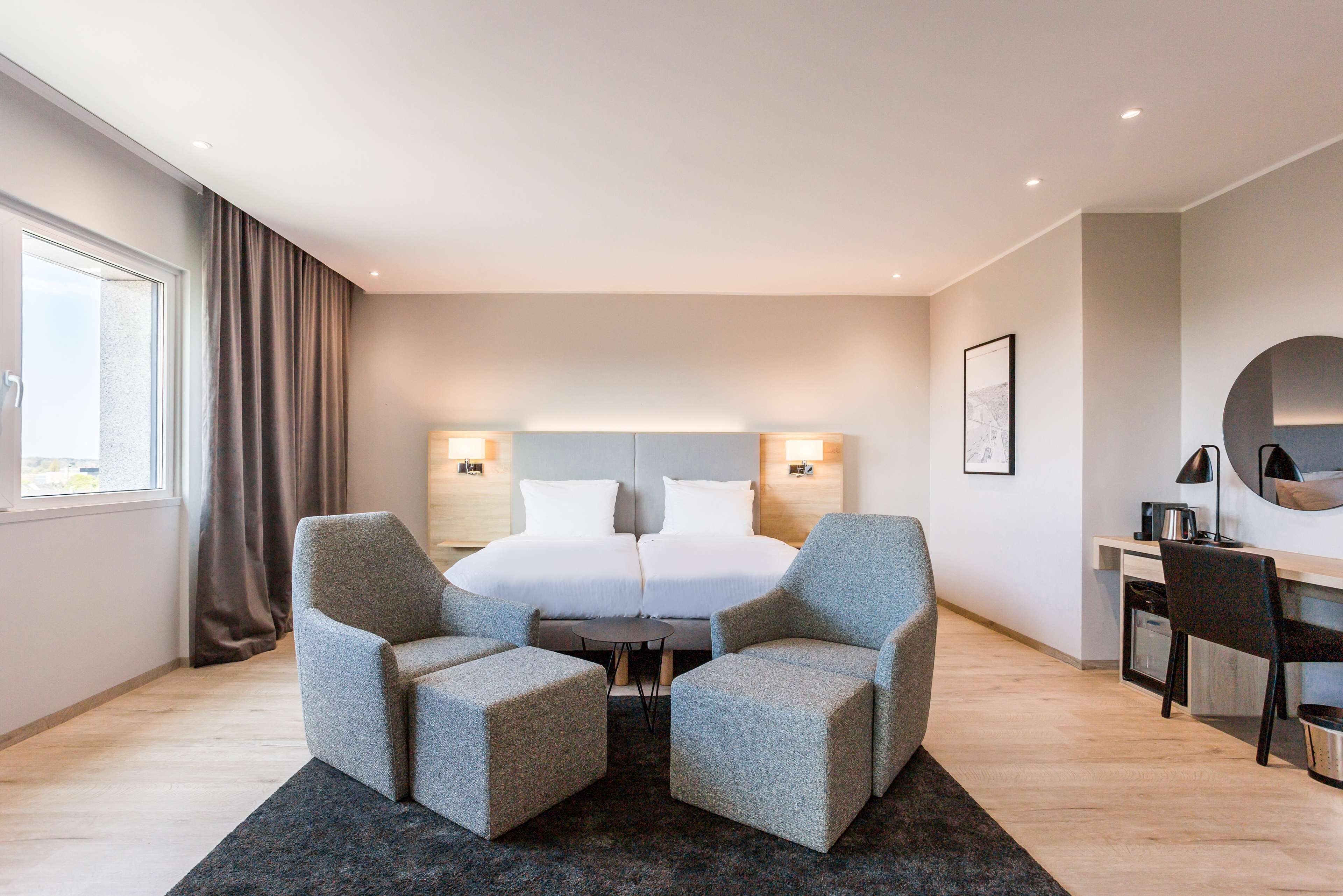 Una habitación de hotel moderna con una cama grande, dos sillones, una mesita, un escritorio con silla, un espejo redondo y una ventana con cortinas.
