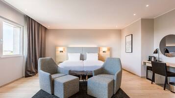 Una habitación de hotel moderna con una cama grande, dos sillones, una mesita, un escritorio con silla, un espejo redondo y una ventana con cortinas.