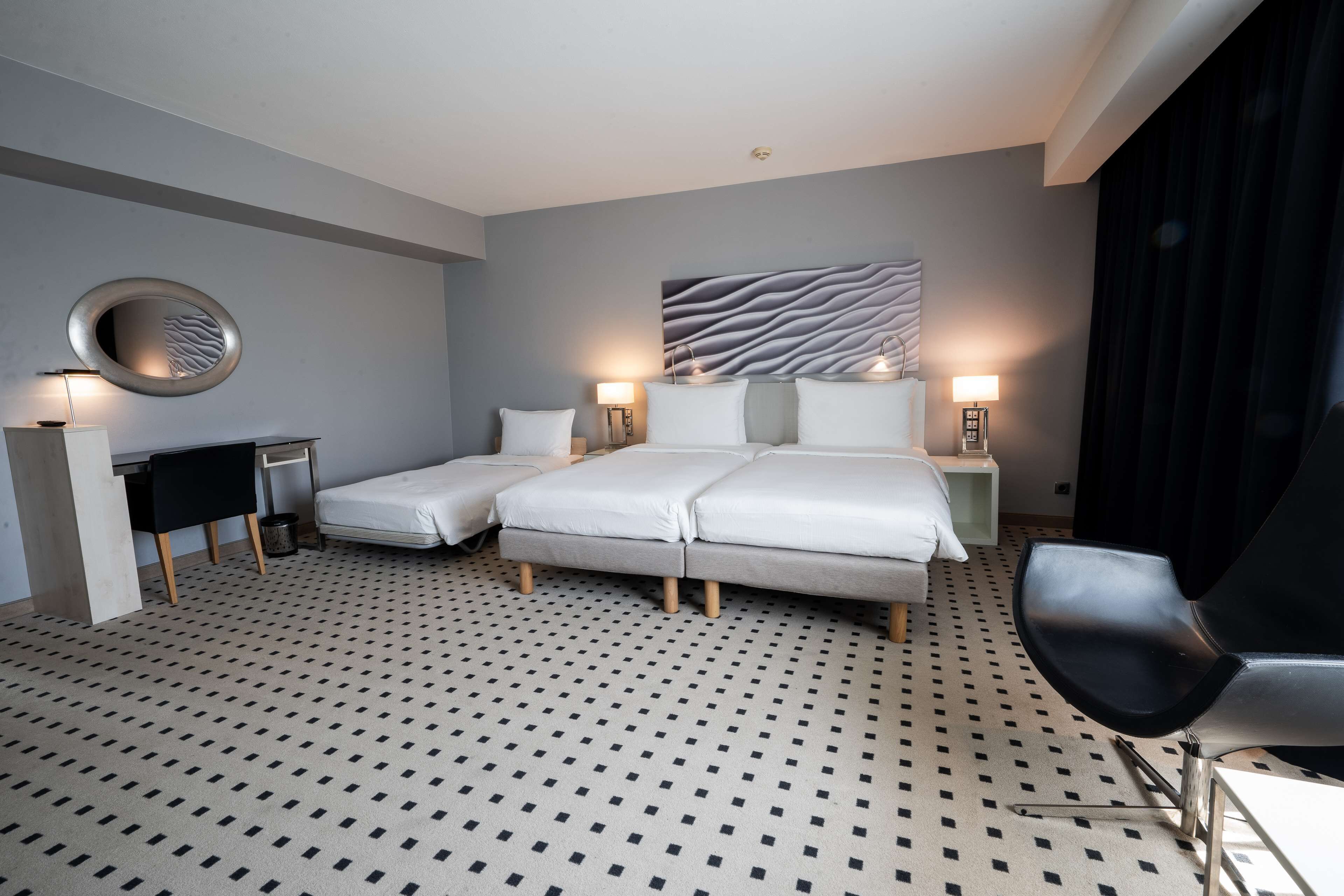 A hotel room with a bed, two bedside tables, a chair, a desk, a mirror, and a patterned carpet.