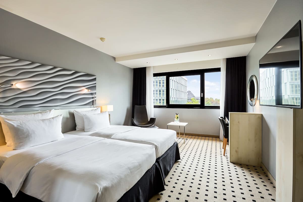 A hotel room with two beds, a modern headboard, a small table, a chair, a large window, and a patterned floor.