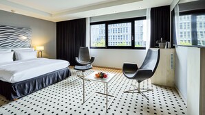 A modern hotel room with a large bed, two chairs, a small table with fruit, and large windows with a city view.