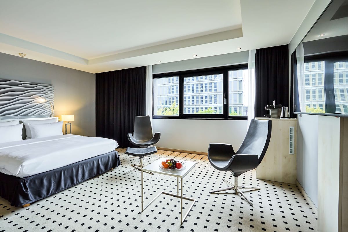 A modern hotel room with a large bed, two chairs, a small table with fruit, and large windows with a city view.
