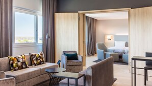 A modern hotel room with a sofa, armchair, and a bed. There is a dining table and a small table with a chair.