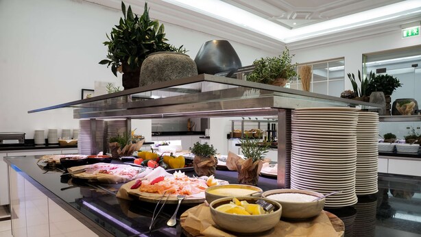 A buffet setup with various food items, a stack of plates, and decorative plants.