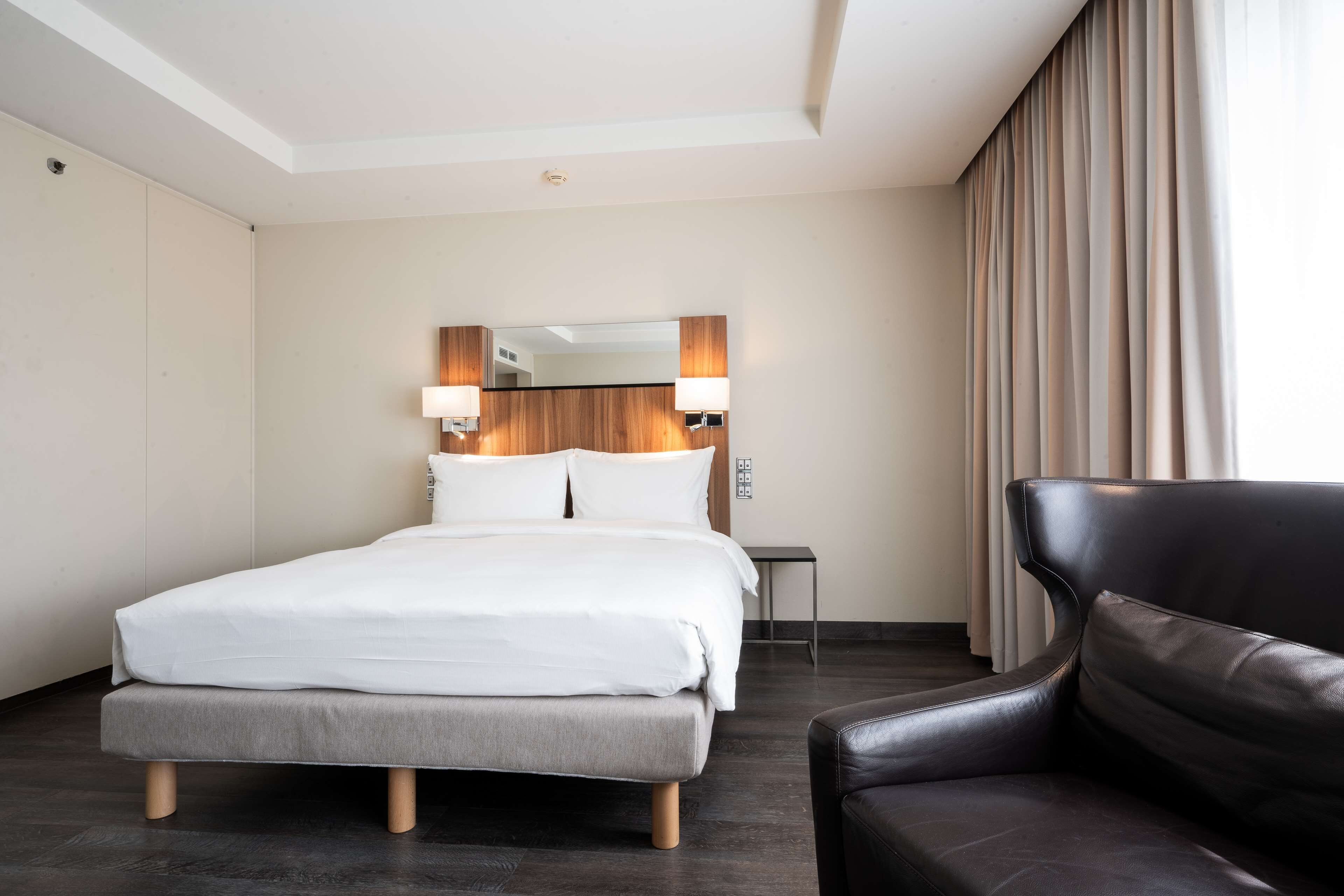 A hotel room with a bed, a chair, a small table, and a mirror with wall-mounted lamps.