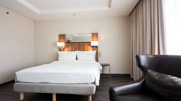 A hotel room with a bed, a chair, a small table, and a mirror with wall-mounted lamps.