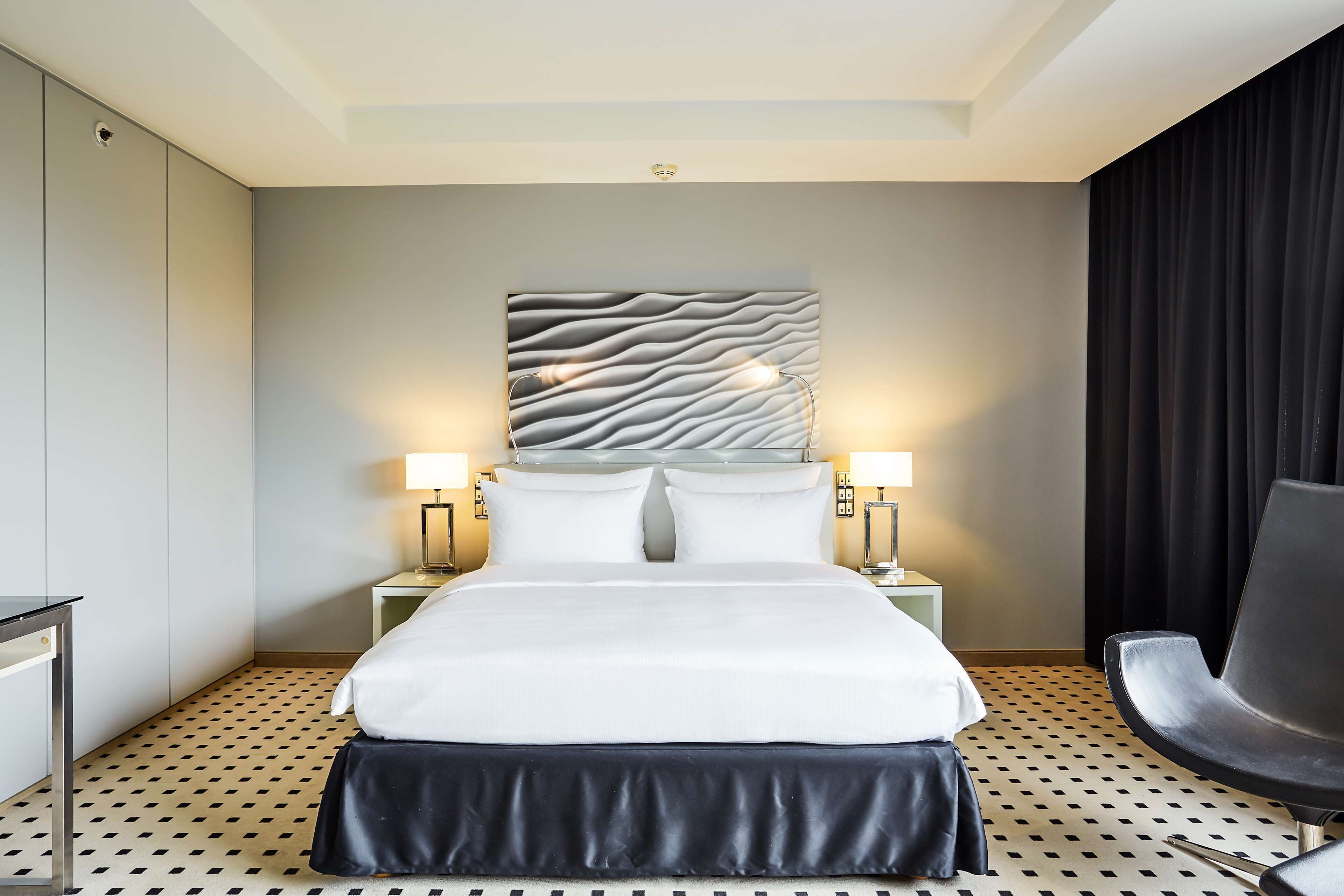 Una habitación de hotel con una cama, dos lámparas de noche, una silla negra y un suelo estampado.