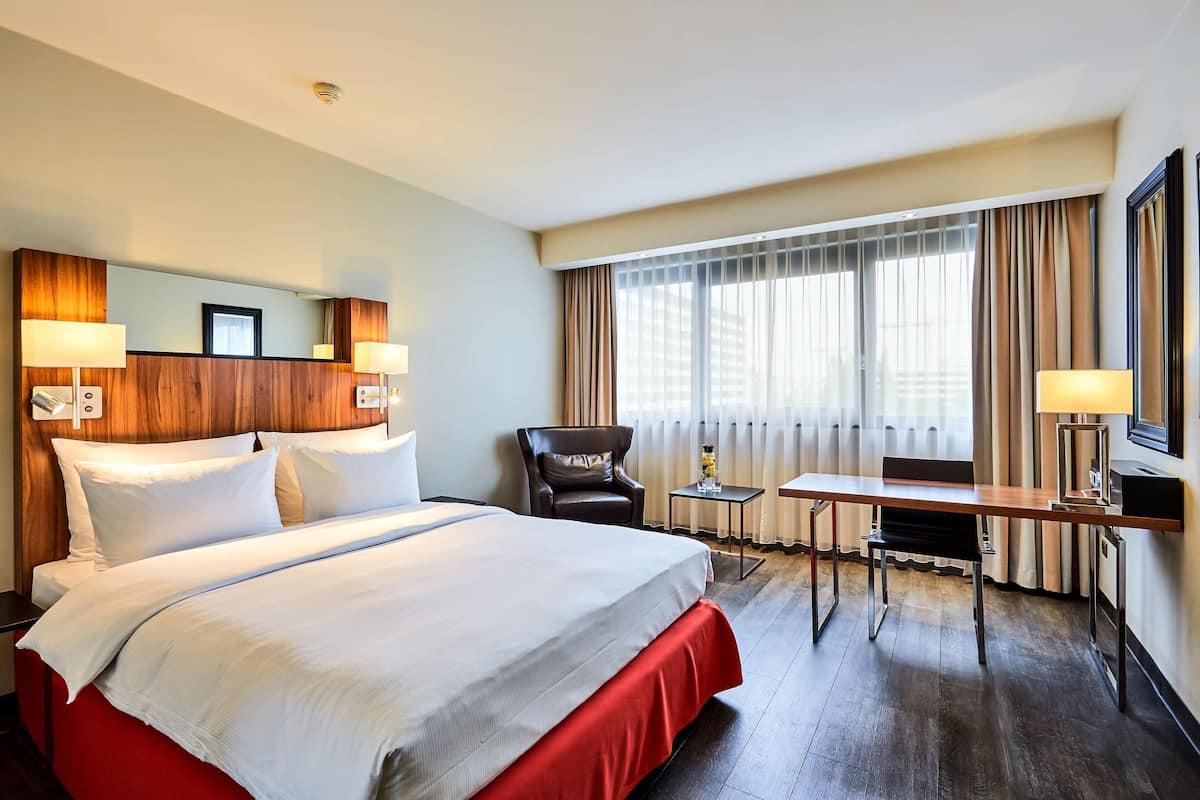 A hotel room with a large bed, a desk, a chair, a small table, and a large window with curtains.