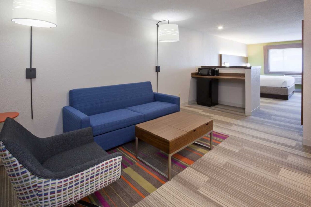 A modern hotel room with a blue sofa, a small wooden table, a colorful striped rug, a patterned chair, and a kitchenette area.