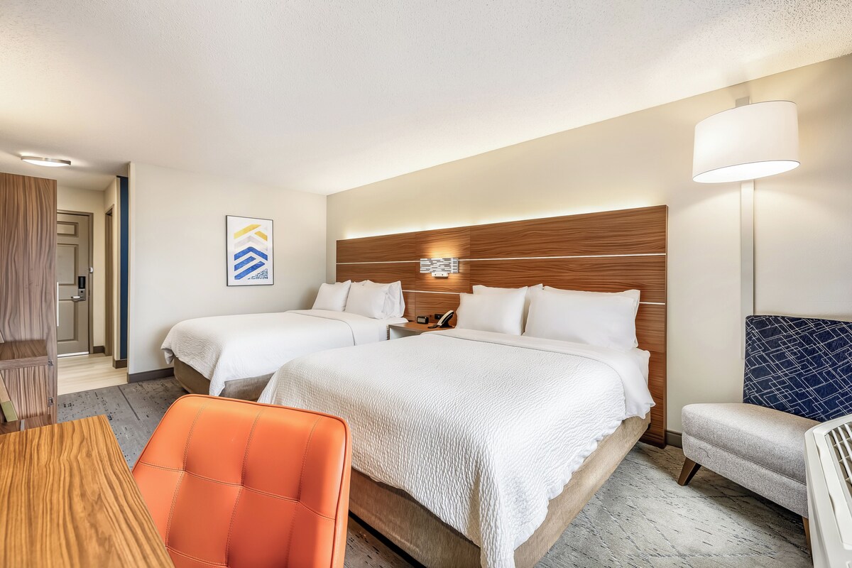 A hotel room with two beds, a desk, a chair, and a lamp.